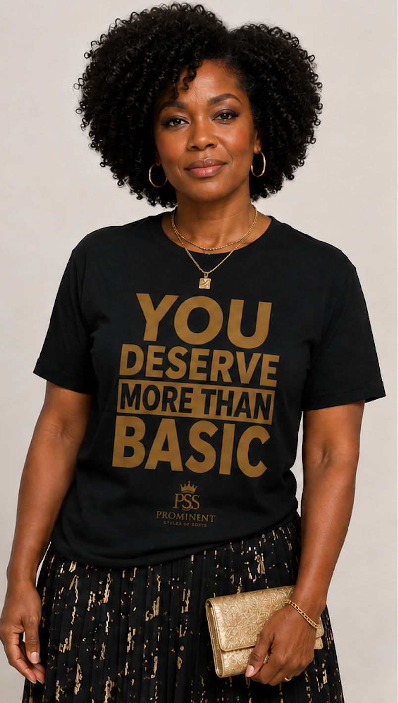 A woman with curly black hair wearing a black graphic t-shirt with gold lettering that reads 'YOU DESERVE MORE THAN BASIC'. She is holding a gold clutch and wearing a black and gold patterned skirt, set against a plain light beige background.